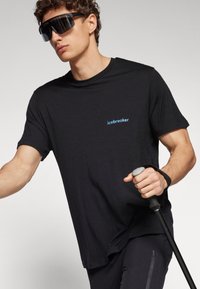 Black short-sleeve T-shirt made of soft fabric with a subtle blue "icebreaker" logo on the left chest; paired with trekking poles.