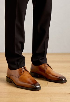 Braune Lederhalbschuhe getragen mit schwarzen maßgeschneiderten Hosen auf lichtfarbenem Holzboden.