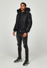 Giubbotto imbottito nero con cappuccio in pelliccia sintetica, motivo trapuntato sulle spalle, abbinato a jeans neri strappati e scarpe sportive nere.