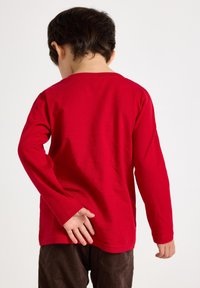 Red long-sleeve shirt made of cotton, featuring a crew neck and a relaxed fit. The child is wearing brown corduroy pants.