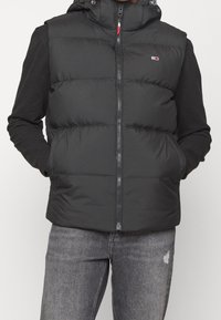Gilet piumino nero con cappuccio, chiusura zip e due tasche laterali. Esterno morbido, design trapuntato, indossato sopra una maglietta a maniche lunghe scura.