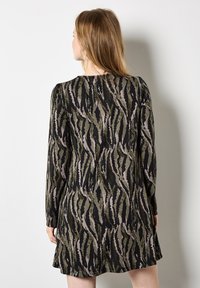 Donna con capelli lunghi che indossa un abito a maniche lunghe con un motivo astratto in nero, verde e beige, in piedi contro un muro bianco, rivolta verso l'esterno.