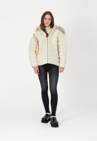 Chaqueta de plumas en color crema con diseño de acolchado horizontal y capucha forrada de piel. Combinada con jeans oscuros y zapatos de plataforma negros, de pie.