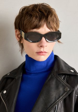 Sorte solbriller med runde kanter og tonede linser. Har guldaccent hardware på skinnerne. Båret med en blå ribbed højhalset trøje og læderjakke.