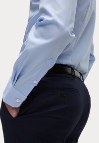 Helles blaues Hemd mit Knopfmanschetten, neben dunkelblauen Hosen mit geknöpftem Gesäßtasche präsentiert. Glattes Gewebe mit maßgeschneidertem Schnitt.