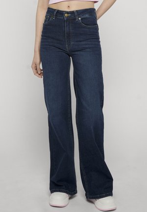 Femme portant un jean large taille haute bleu foncé et des baskets blanches, debout sur un fond clair uni.