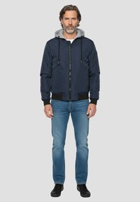 Marineblaue Bomberjacke mit grauer Kapuze, Frontreißverschluss, elastischen Bündchen und Bund; kombiniert mit hellblauen Jeans und schwarzen Schuhen.