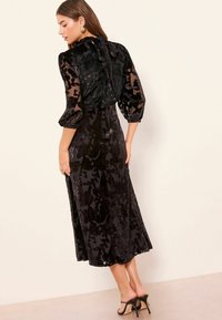 Robe en velours noir à motif floral, manches longues transparentes et détail dos en forme de goutte. La jupe offre une silhouette fluide.