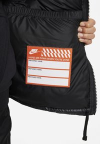 Svart puffjacka med inre som visar en stänkt orange etikett med randig kant och text för föredraget namn, fäst vid foder.