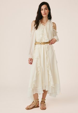 Kobieta stojąca w białej, marszczonej sukience z odsłoniętymi ramionami, z paskiem ozdobionym złotymi ćwiekami i dopasowanymi złotymi sandałami gladiator.