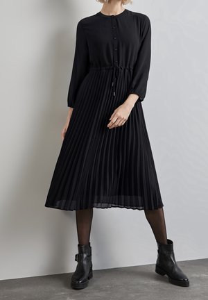 Woman wearing a black long-sleeve buttoned top with drawstring waist and pleated midi skirt, black tights, and black ankle boots.