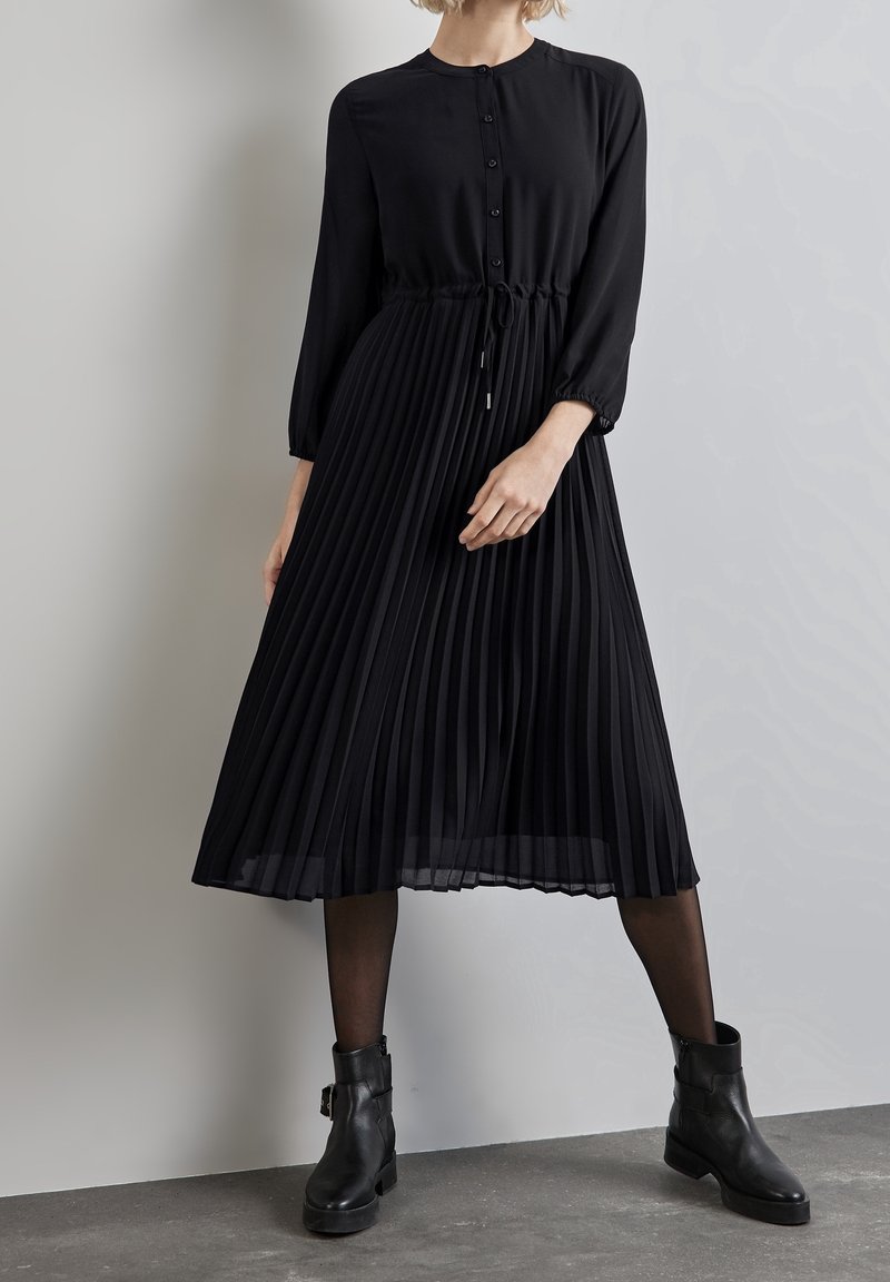Femme portant un haut noir à manches longues boutonné avec une taille à cordon et une jupe midi plissée, des collants noirs et des bottines noires.