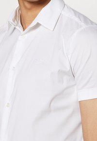 Kortärmad vit skjorta i mjukt tyg, med knäppning framtill och en diskret broderad logo på bröstet, samt klassisk krage.