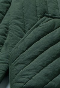 Tessuto verde trapuntato con una texture morbida. Il materiale presenta cuciture diagonali che creano un motivo di sezioni rialzate e piatte.