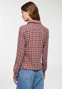 Donna di spalle che indossa una camicia a maniche lunghe a quadri rossi, neri e bianchi con jeans in denim blu, su uno sfondo bianco semplice.
