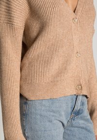 Tan ribbat cardigan med V-ringning, med tre knappar fram och avslappnad passform, kombinerad med ljusblå jeans.