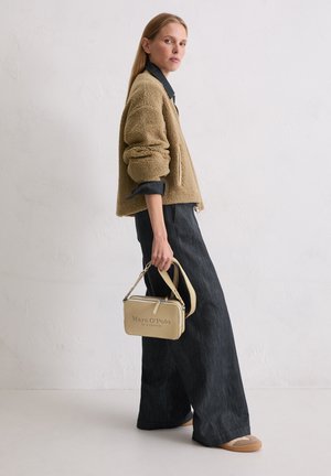 Frau steht im Seitenprofil, trägt eine beige Fleecejacke, schwarze weite Hose, hält eine beigefarbene Marc O'Polo Handtasche seitlich.