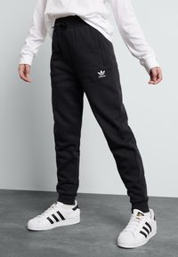 Schwarze Fleece-Jogginghosen mit einem elastischen Bund, Seitentaschen und einem kleinen weißen Adidas-Logo. Kombiniert mit weißen Adidas-Sneakers mit schwarzen Streifen.