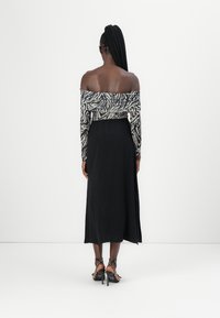Femme aux longues tresses porte un haut zébré sans épaules, une jupe midi noire avec une fente latérale et des sandales noires à talons hauts cloutées, debout de dos.
