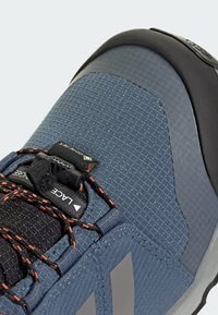 Baskets en tissu technique bleu avec des accents gris, surface texturée, capuchon noir, lacets orange contrastants et un système de laçage avec marque.