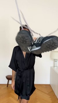 Robe de cetim preto com mangas largas e cintura ajustada, exibindo ténis atléticos pretos com detalhes brancos e solas texturizadas.