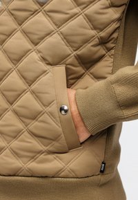 Χέρι μερικώς μέσα στην τσέπη μιας ταμπά καπιτονέ μπουφάν με κουμπί μπροστά, μανίκι και μέση από πλεκτό ύφασμα.