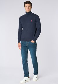 Maglione a collo alto navy con orlo a coste, abbinato a jeans blu e sneakers bianche. Presenta un logo rosso sul petto.