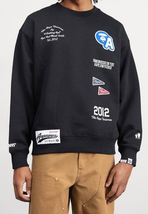 Sudadera negra con texto "Aape Universe" en blanco y azul, logotipos y parches combinados con pantalones color beige en una persona de cintura para arriba.