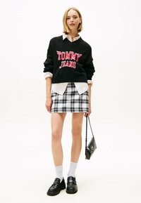 Schwarzer Pullover mit roter Aufschrift "TOMMY JEANS", über einem weißen Hemd getragen. Kombiniert mit einem karierten Minirock und schwarzen Loafers.