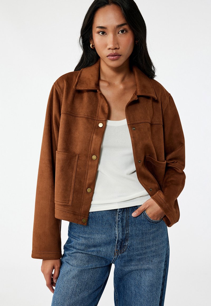 Brun mockaskinnjacka med krage, tryckknappar, framficka och kort design. Bärs över en ljus linne och blå jeans.