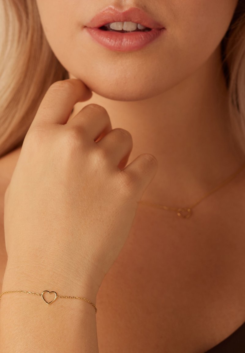 Pulsera de oro con un colgante en forma de corazón, llevada en una mano. Textura de piel suave, diseño minimalista, combinada con un collar de oro a juego.