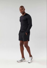 Zwarte lange mouwen sporttop met een getailleerd ontwerp, gecombineerd met zwarte shorts. Model draagt witte en grijze hardloopschoenen met gele accenten.