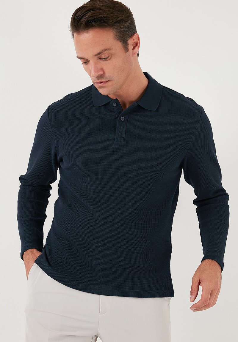 Polo de manga larga en color marino con un tejido texturizado, que presenta un cuello clásico y una botonera de tres botones, combinado con pantalones de color claro.