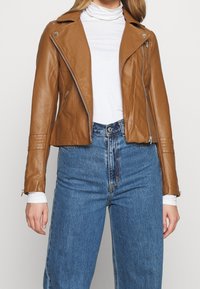 Brun læder-motorcykeljakke med tætsiddende design, asymmetrisk lynlås og lynlåse ved ærmerne, båret over en hvid rullekrave og blå jeans.