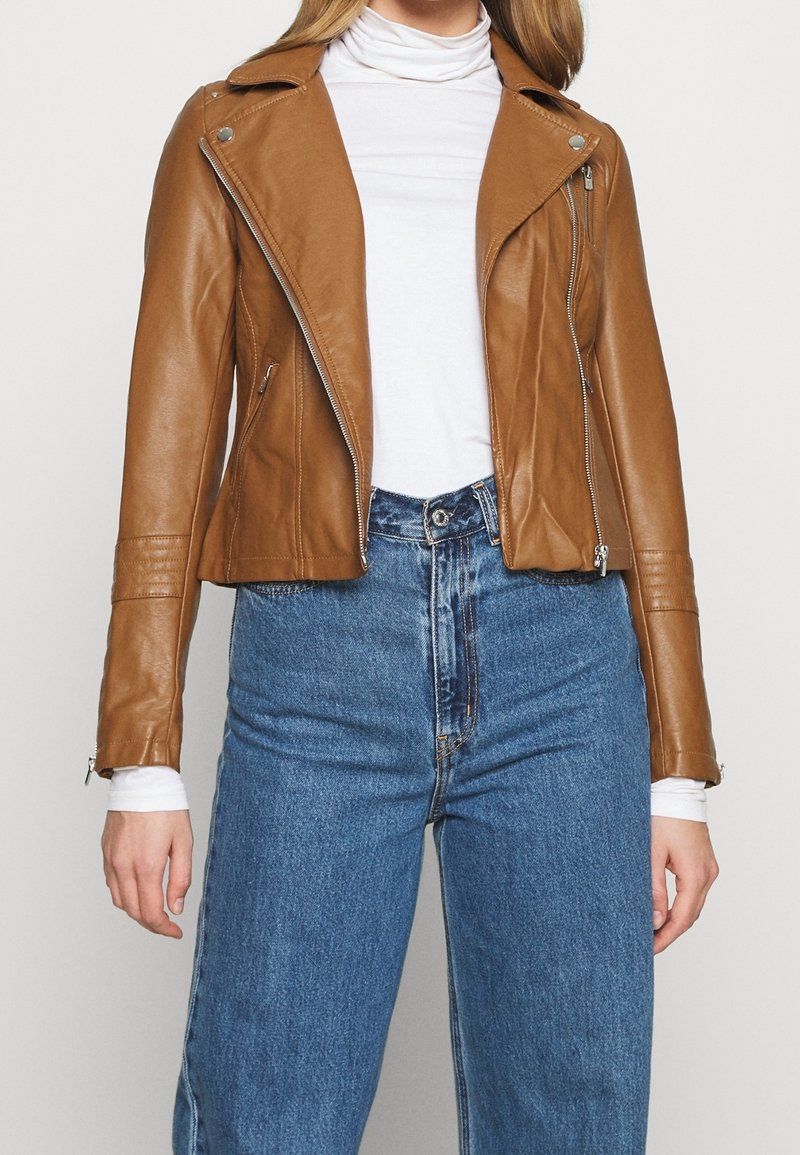 Brązowa skórzana kurtka motocyklowa o dopasowanym kroju, z asymetrycznym zamkiem błyskawicznym i zamkami przy mankietach, noszona na białym golfie i niebieskich dżinsach.