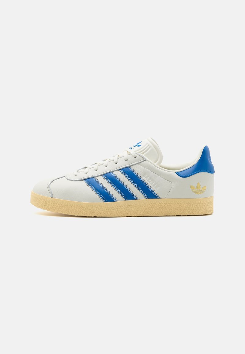 Zapatilla de piel blanca con detalles azules y tres rayas azules. Suela de goma, puntera texturizada de color marrón y logotipo dorado en el talón.