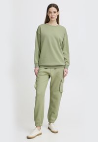 Felpa verde in cotone con polsini a coste e collo rotondo, abbinata a pantaloni cargo coordinati con tasche laterali e caviglie elastiche.