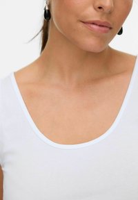 Haut blanc à manches courtes et décolleté large, avec une texture lisse semblable au coton, accompagné d'une boucle d'oreille pendante.