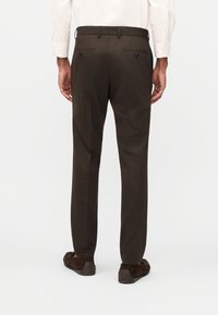 Άνδρας που φοράει σκούρο καφέ ραμμένο παντελόνι, λευκό πουκάμισο και σκούρα καφέ μοκασίνια, στέκεται με την πλάτη προς το μέρος μας σε απλό φόντο.