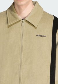 Green corduroy jacket with a zip front, large collar, and black vertical stripe. Features an embroidered black Adidas logo on the chest.