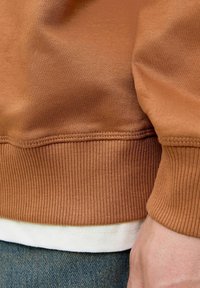 Felpa marrone in tessuto a costine, con orlo bianco e cuciture tono su tono. Primo piano della manica che mostra dettagli della texture e della vestibilità su jeans.