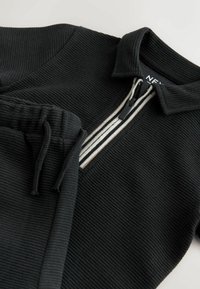 Zwarte geribbelde pullover met halve rits en gestreepte rand aan de kraag, gecombineerd met bijpassende geribbelde broek met trekkoord in de taille.