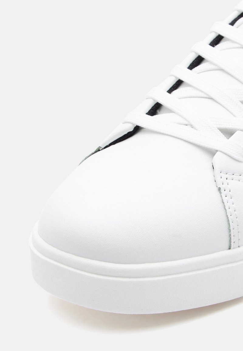 Zapatilla de cuero blanco con punta redondeada, suela plana y detalles de encaje intrincados. Textura suave con acentos mínimos.