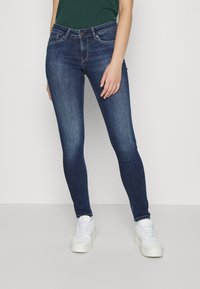 Jeans skinny azul-escuro com uma textura denim suave, apresentando bolsos frontais, passantes para cinto e fecho de botão, combinados com sapatilhas brancas.