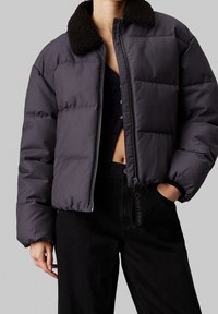 Kobieta w ciemnofiołkowej kurtce puchowej z czarnym kołnierzem z owczej skóry, częściowo zapięta nad czarnym topem z krótkim rękawem i czarnymi spodniami z wysokim stanem.