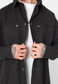 Veste noire à boutons sur le devant avec deux poches poitrine, fabriquée en tissu texturé. Elle présente des boutons-pression et un logo de marque subtil sur la poitrine.