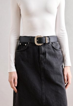 Jupe en denim noir à taille haute, associée à un haut blanc à manches longues. Une ceinture en cuir noir arbore une boucle en argent texturée et torsadée.