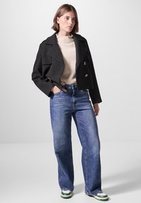 Avkortad svart dubbelknäppt kavaj, ljusbeige polotröja, blå vida jeans och sneakers med gröna detaljer. Sätt att kombinera släta texturer och klassisk design.
