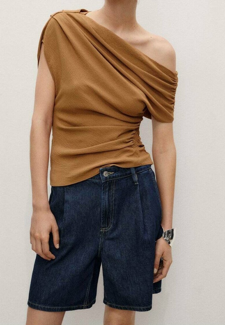 Top marrone drappeggiato con design monospalla, abbinato a pantaloncini di denim blu scuro. Il top presenta dettagli arricciati sul lato. Orologio d'argento al polso.