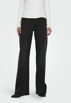 Kvinde iført sorte jeans med vide ben, sorte sko og en hvid top med lange ærmer, står med hænderne afslappet langs siden.